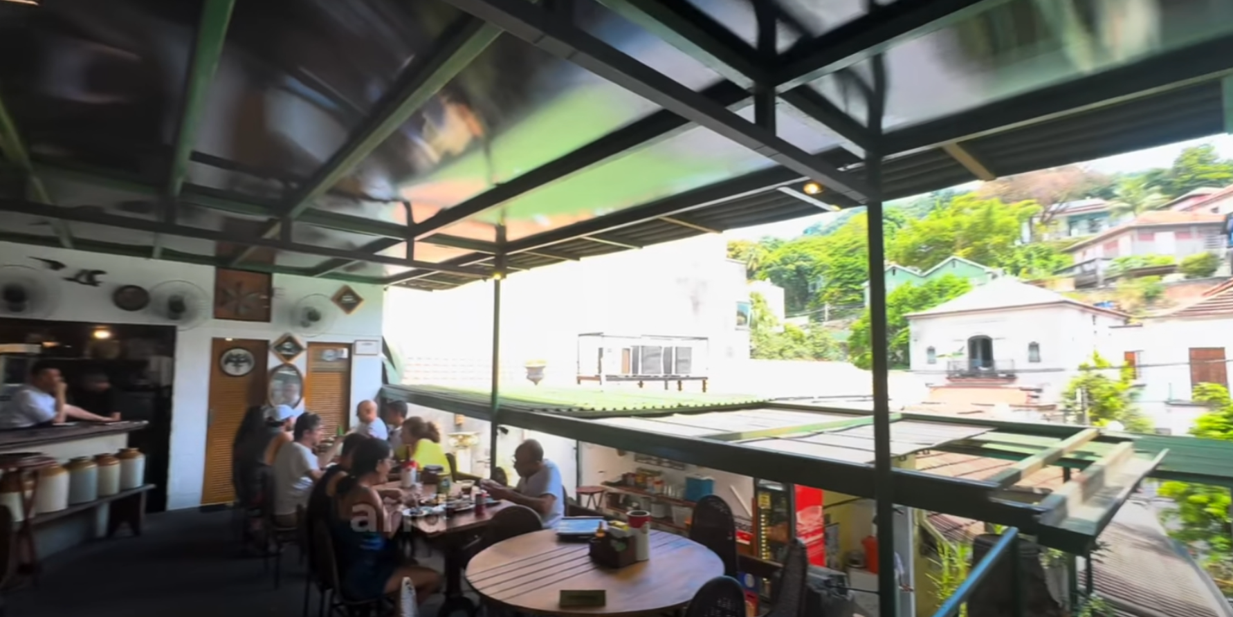 People dining and chatting at an open-air restaurant with a scenic hillside view. Rustic decor, relaxed atmosphere, sunny day.