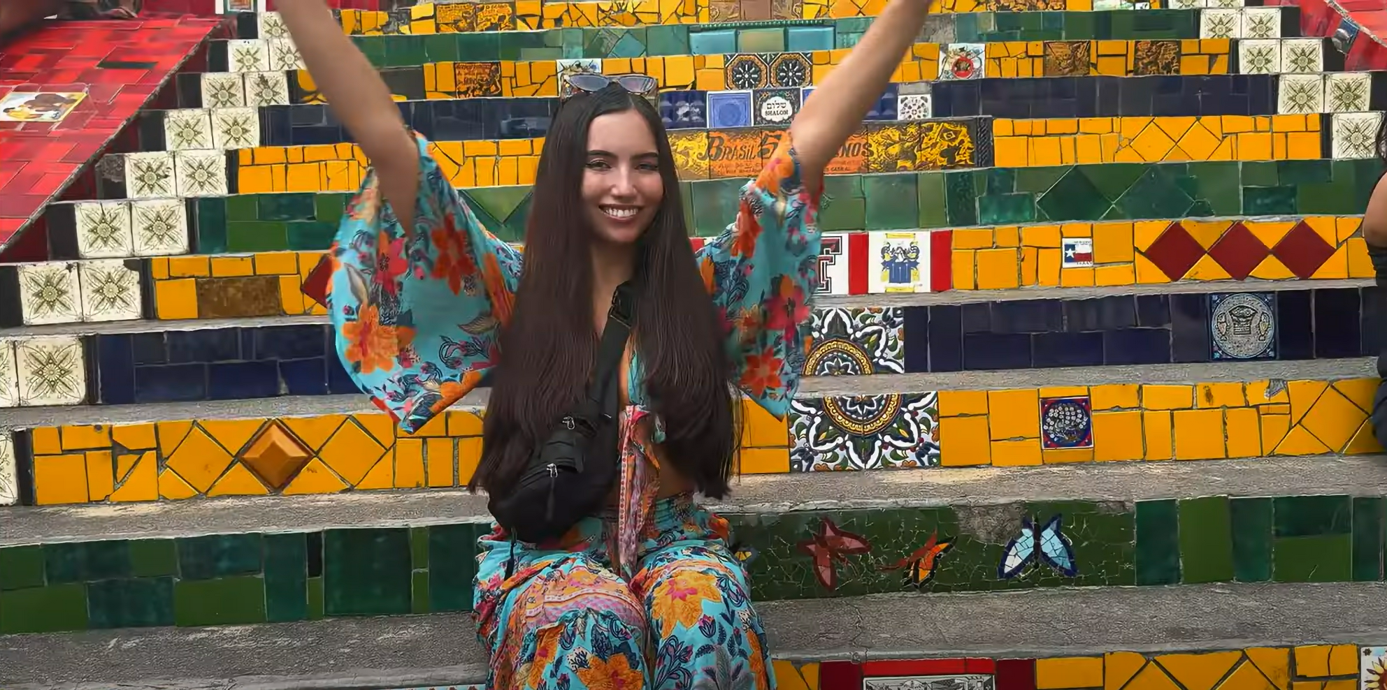 Woman smiles with arms raised, sitting on colorful tiled stairs. She's wearing a floral dress and sunglasses, exuding a joyful mood.