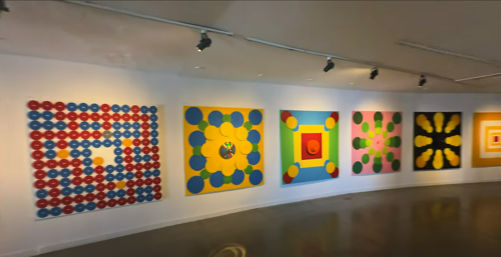 Art gallery with colorful geometric paintings on a curved white wall, featuring circles, squares, and vibrant patterns under spotlights.
