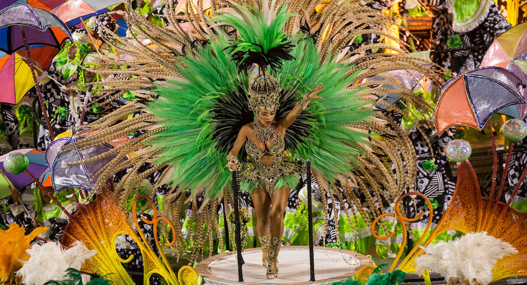 Carnival dancer in vibrant green and gold feathered costume stands on a float. Colorful umbrellas and patterns create a festive backdrop.