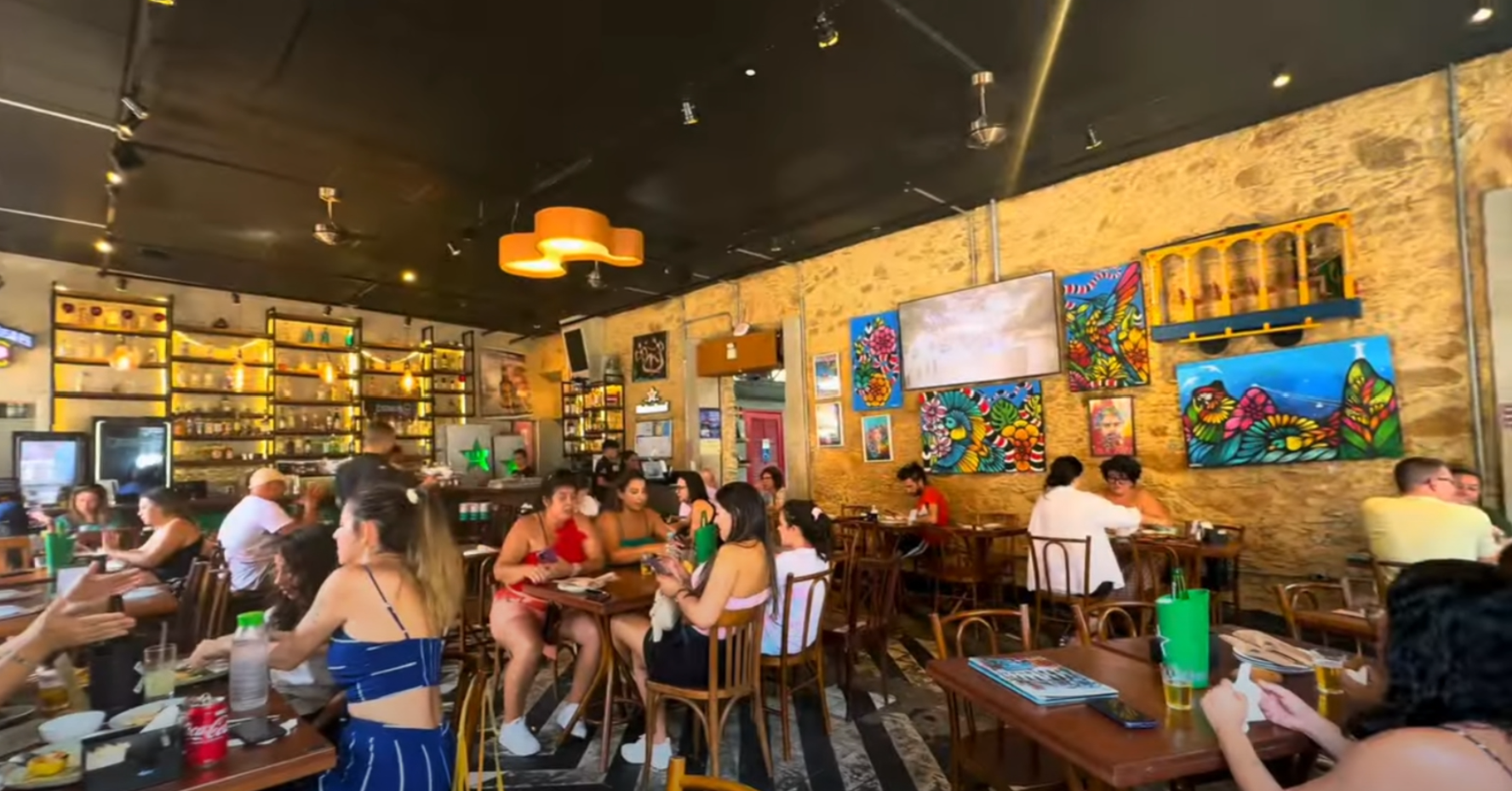 People dining in a vibrant restaurant with colorful artwork on stone walls. Warm lighting, lively atmosphere, and drinks on tables.
