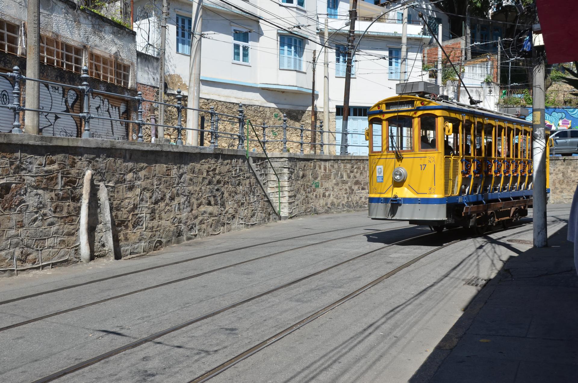 A bright yellow tram travels along the street next to stone walls and graffiti-covered buildings under a clear blue sky.
