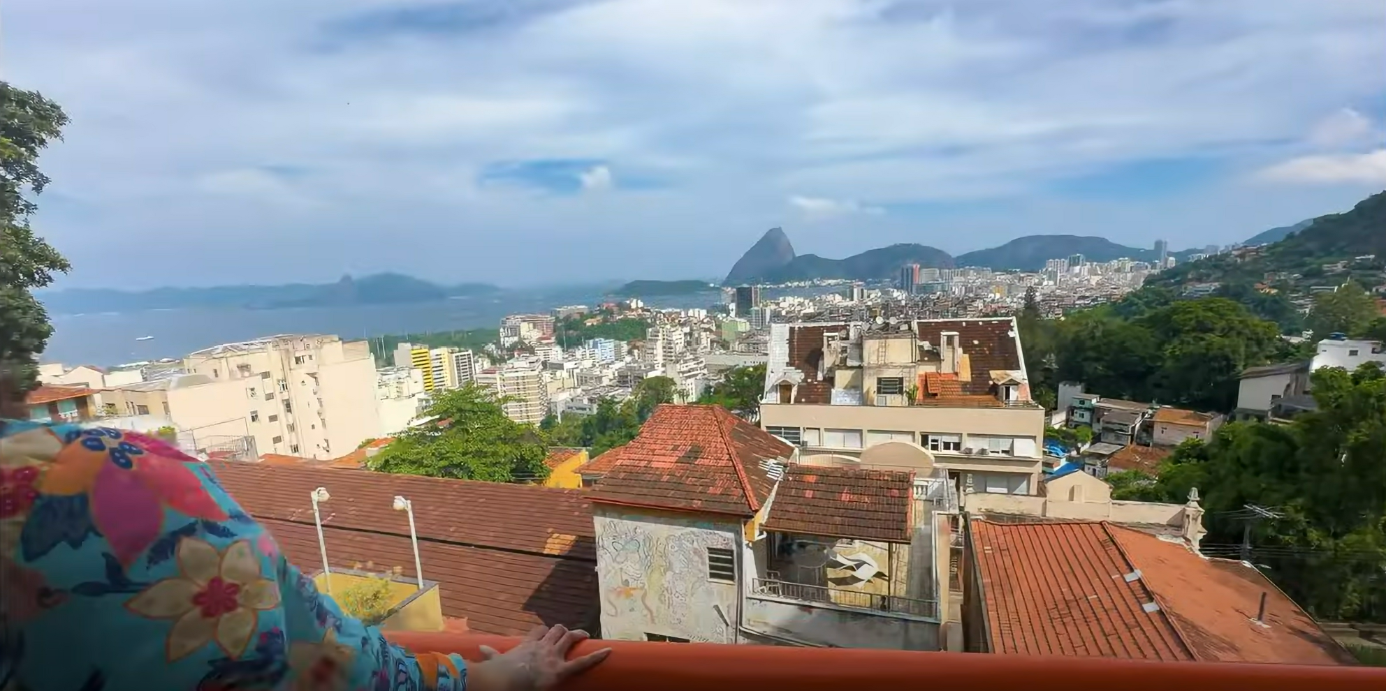 Colorful figure gazing over Rio de Janeiro's skyline with Sugarloaf Mountain in the distance, rooftops and greenery under a bright sky.