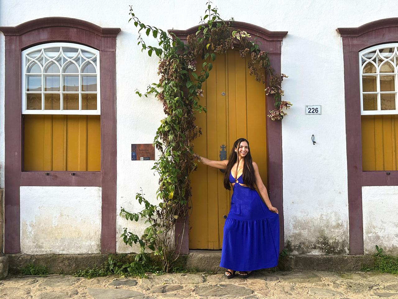 Woman in a blue dress poses cheerfully by a yellow door. White wall with brown accents, climbing plants, and window panes create a charming setting.