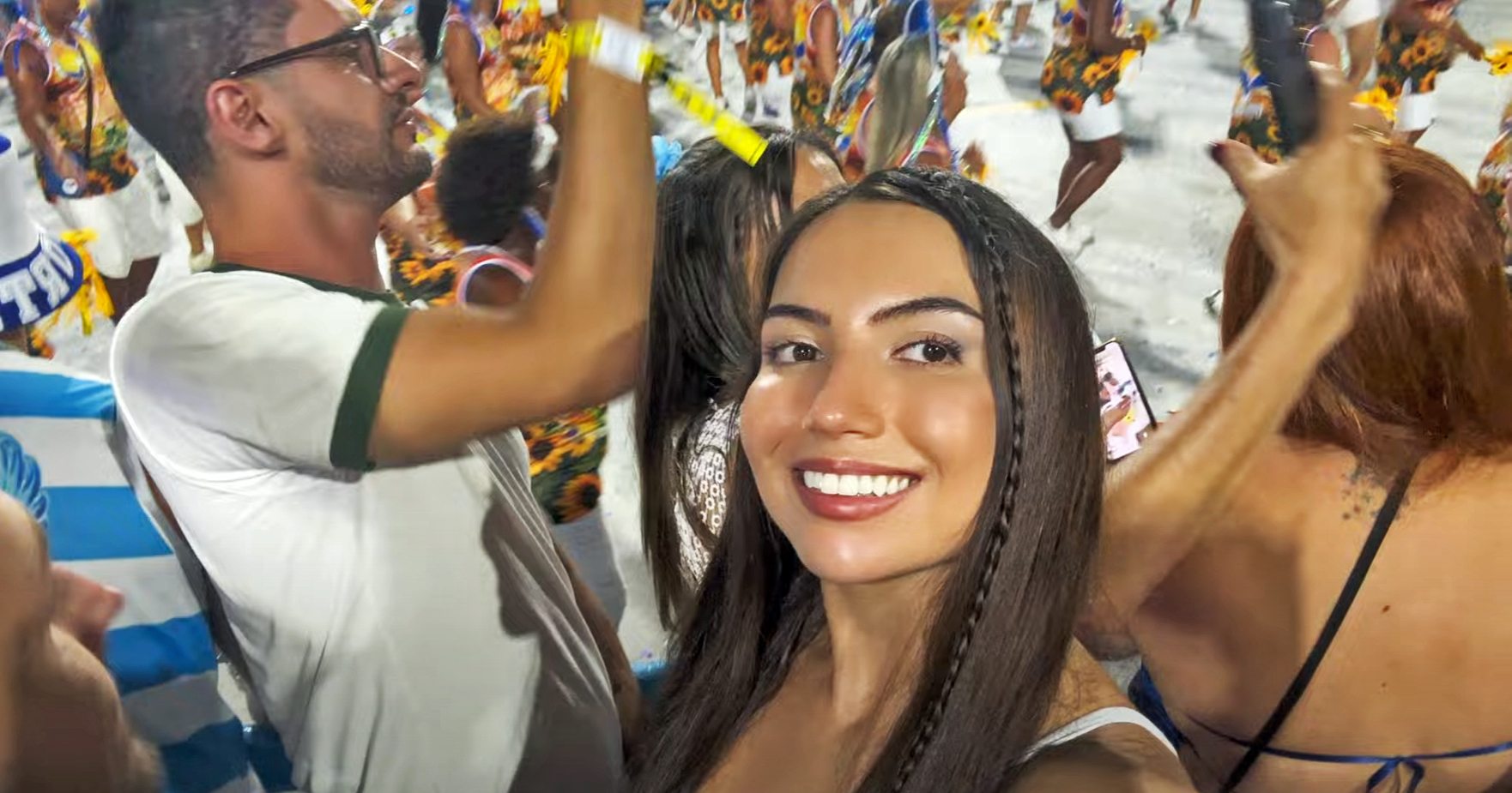 Smiling woman taking selfie at a lively parade with colorful dancers and joyful crowd in background. Vibrant, festive atmosphere.