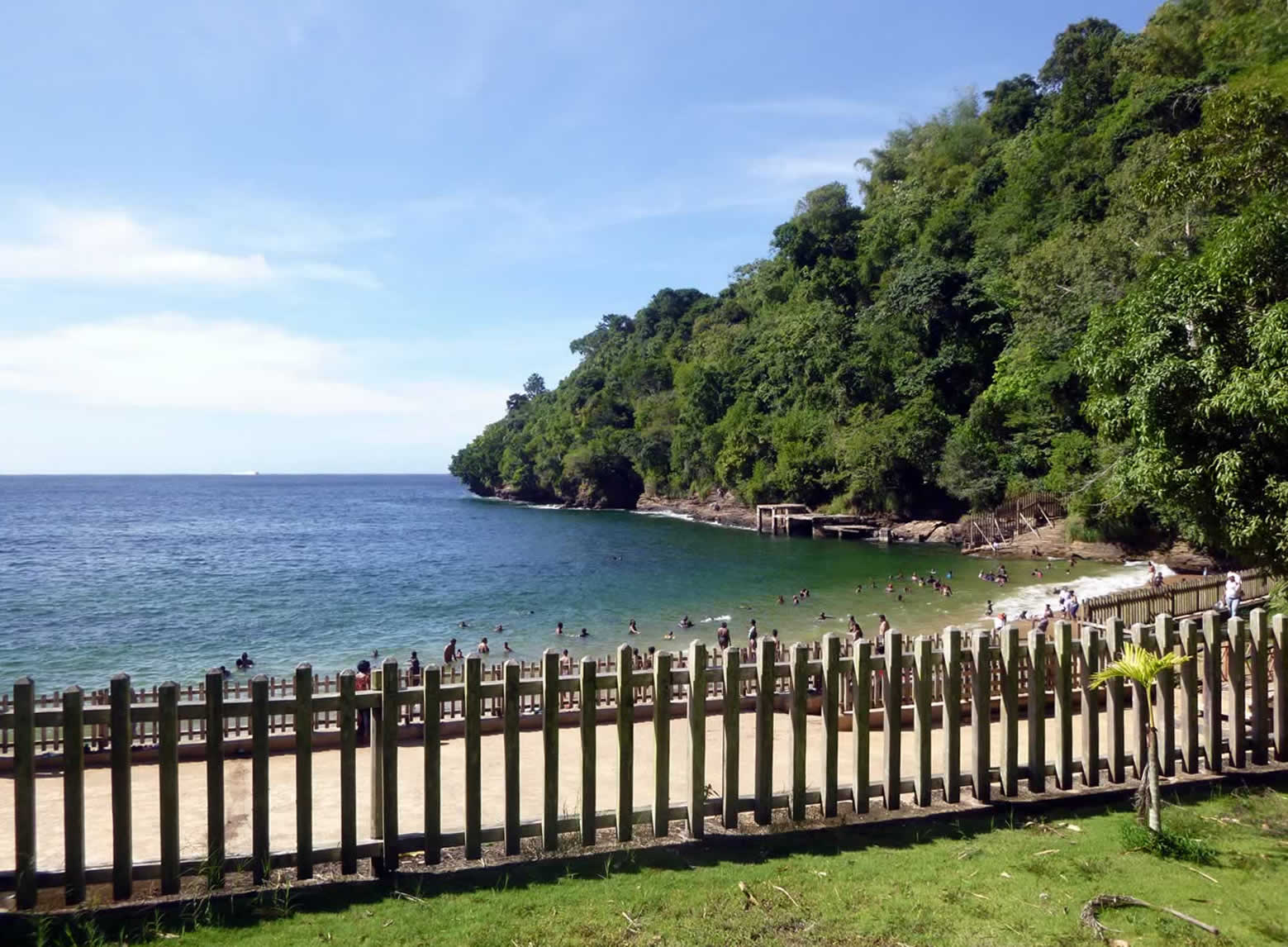 People swim in a sunny bay with blue water and green hills. A wooden fence lines the beach, creating a lively and relaxing atmosphere.